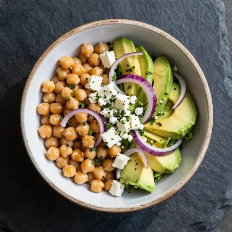 A vibrant chickpea salad with feta cheese, avocado slices, and fresh greens served in a white bowl.
